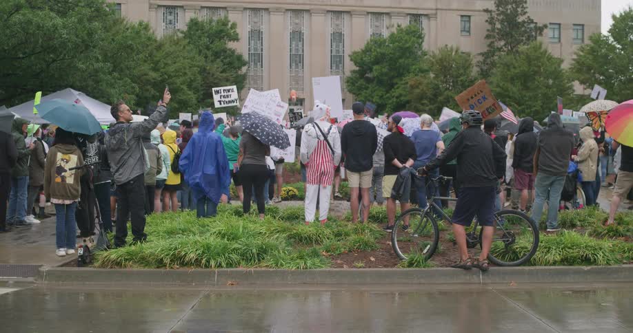 Protesters in rain gear hold signs outside municipal building on damp day
