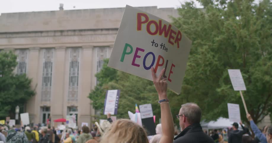 The video shows a protest or demonstration in a public space focusing on a person holding a handmade sign high above the crowd the sign reads power to the people in large colorful lettering with a classical government or institutional building in th