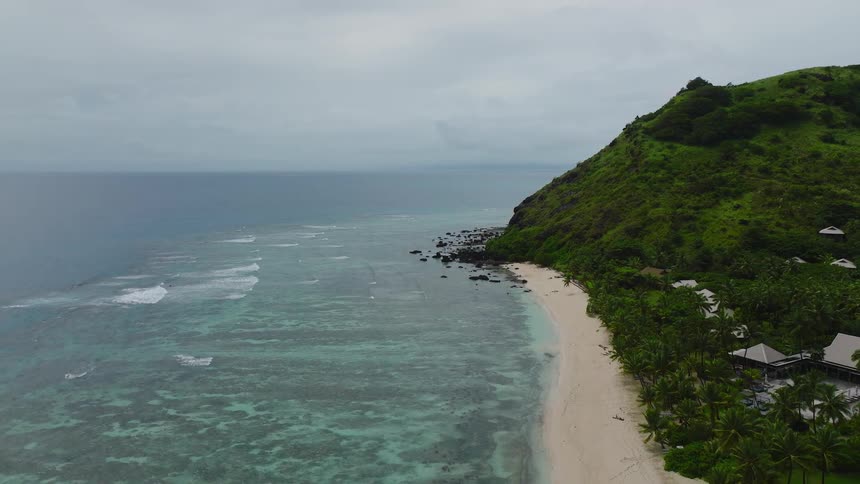 An aerial view shows a lush green hill sloping to a white sand beach and turquoise ocean under an overcast sky