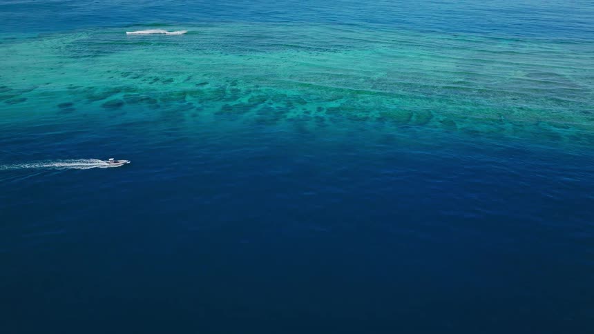 An aerial view shows deep dark blue ocean contrasting with a large vibrant turquoise and light blue shallow coral reef area near the top of the framea small white speedboat cuts across the deep blue water in the foreground leaving a distinct white w