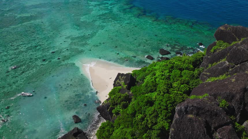 The scene features a dramatic tropical coast where steep dark jagged cliffs covered in brilliant green jungle meet a small sheltered crescent of white sand beach