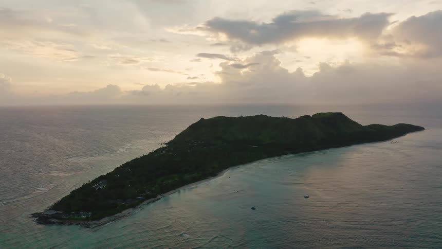 An elevated wide shot captures a long dark heavily vegetated tropical island rising from the ocean under a dramatic cloudy sky at sunsetthe island is surrounded by clear light turquoise shallow water which transitions to the deep darker blue of t