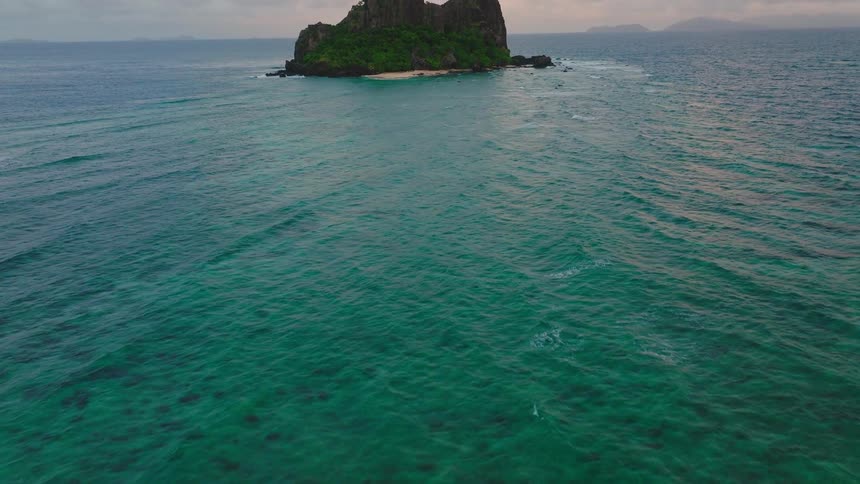 An aerial shot begins close over rippling turquoise ocean water revealing clear shallow depths and then pulls back and rises to unveil a steep dark green rocky islet