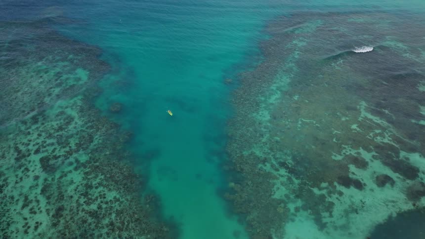 An overhead drone shot captures a coral reef system in clear turquoise tropical water with a person paddling a yellow kayak through a deeper sandy channel that cuts through the dark reef structure