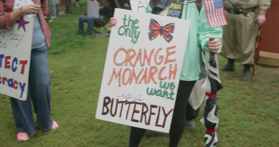 Woman standing up holding a protesting sign next to another person with a sign and other people around outdoors during day time