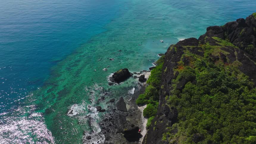 Lush cliffside descends into turquoise ocean with gentle waves breaking on rocks