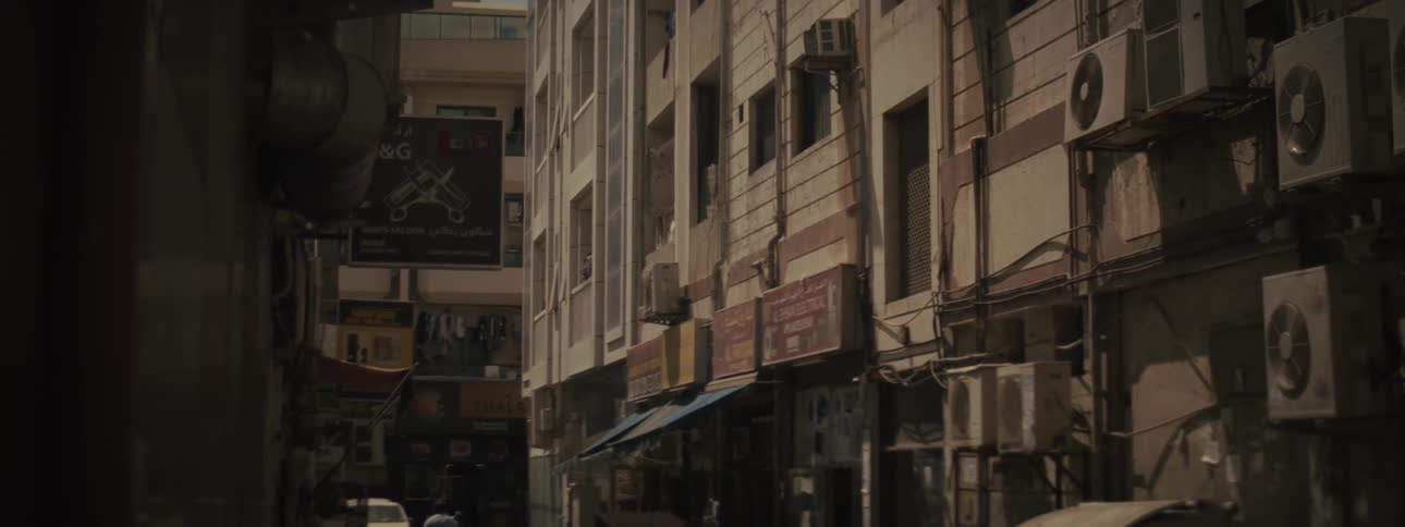 A steady midrange shot captures a narrow alleyway or side street lined with old multistory urban buildings featuring numerous window air conditioning units and faded commercial signs