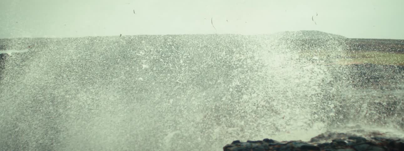Powerful wave crashes against dark jagged rocks spraying water under cloudy sky