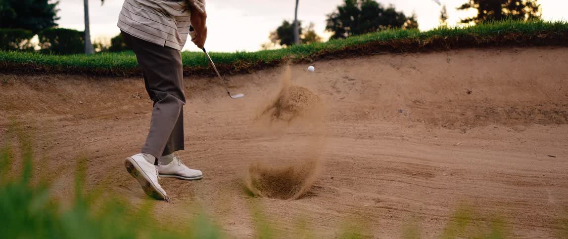 A golfer dressed in slacks and white golf shoes takes a swing at a golf ball situated in a sand bunker causing a visible explosion of sand as the ball is launched out of the trap