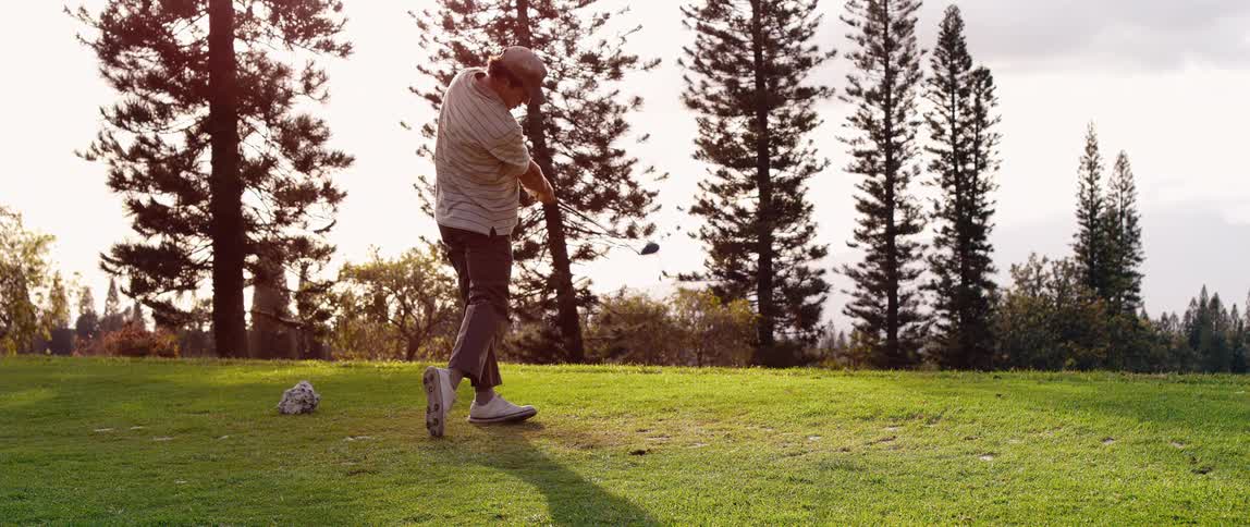 A golfer is shown swinging a driver hitting a golf ball off the tee on a sunny day he is wearing a striped shirt and a cap and the background features tall pine trees silhouetted against the bright sky