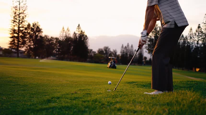 A person wearing a striped shirt and dark trousers shows them standing over a golf ball on the green swinging a golf club to make contact and launch the ball into the air with a golf cart and treelined mountains visible in the distance