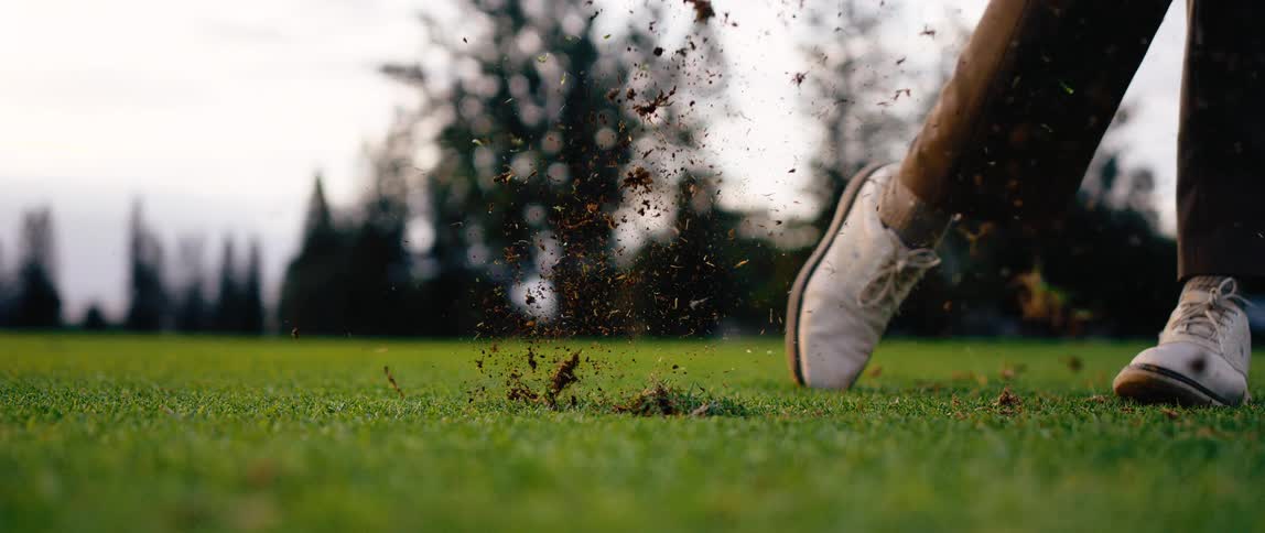 Club strikes ball forcefully sending it airborne as grass and soil scatter from the impact