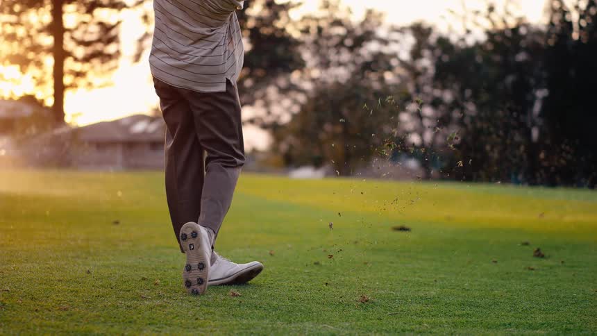 A man on a golf course swings and hits the ball as sprinklers spray across the field with the warm glow of sunset casting light over the scene