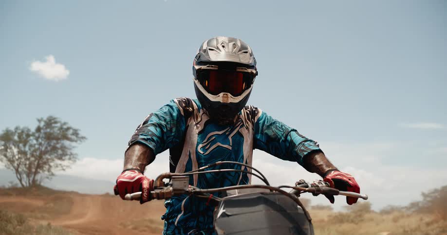 This closeup video filmed from the front shows a motocross rider wearing a helmet and goggles sitting atop a dirt bike on a dusty outdoor track the rider looks slightly to their right toward the end of the clip against a bright clear blue sky