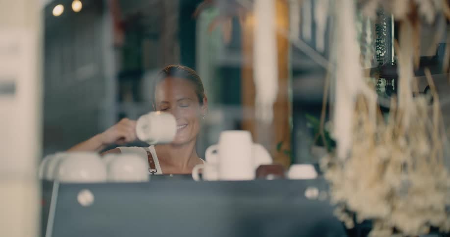 A smiling barista seen through a glass window amidst hanging dried plants holds a white mug near a coffee machine setup