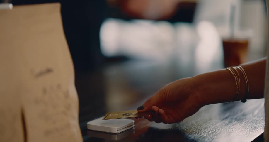 A closeup of a persons hand wearing bracelets inserting a gold credit card into a small square payment reader on a wooden counter the transaction takes place near a brown paper bag suggesting a retail purchase
