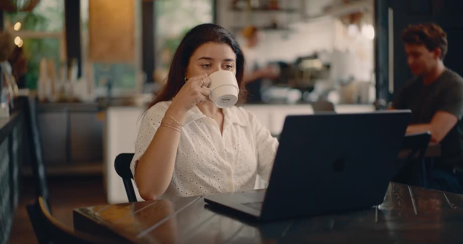 A man and woman are at separate tables in a restaurant looking at their laptops