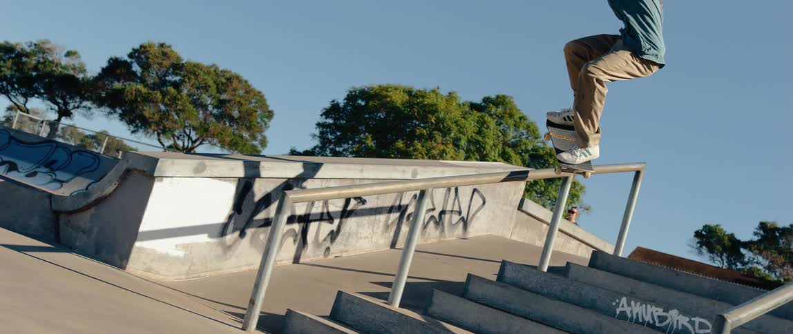 A skateboarder is performing a trick grinding a rail that runs alongside a set of concrete stairs the action takes place at a skate park under a bright blue sky with graffiti visible on the surrounding concrete structures