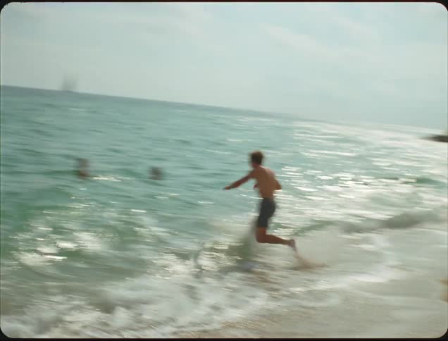 A teenager runs across the sand into the water near other people
