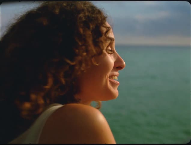 A young woman with curly hair sits at the shoreline at midday smiling as she watches the ocean