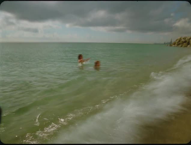 This shaky vintagestyle video features two young girls swimming in clear green ocean water under a partly cloudy sky the sunlight creates a strong sparkling reflection on the waters surface as they play