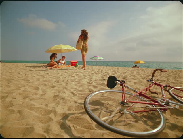 A woman walks away from her bike on a beach toward other people
