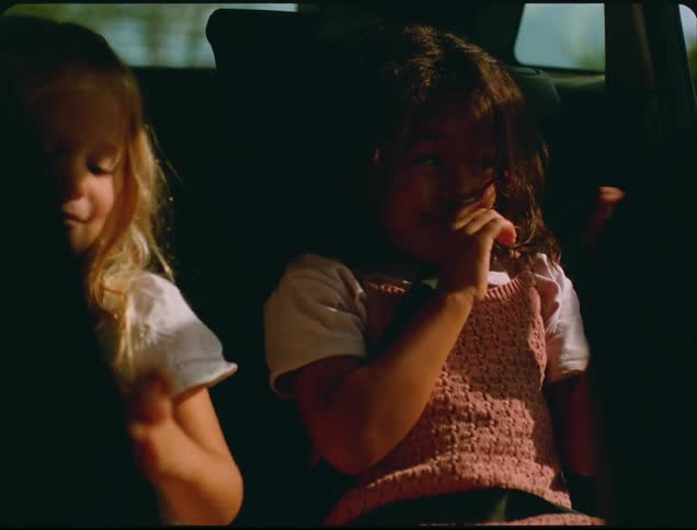 Two young girls in the backseat of a car captures the girl on the left resting her head with her eyes closed while the girl on the right looks around fidgets with her hands and moves her mouth all illuminated by bright light