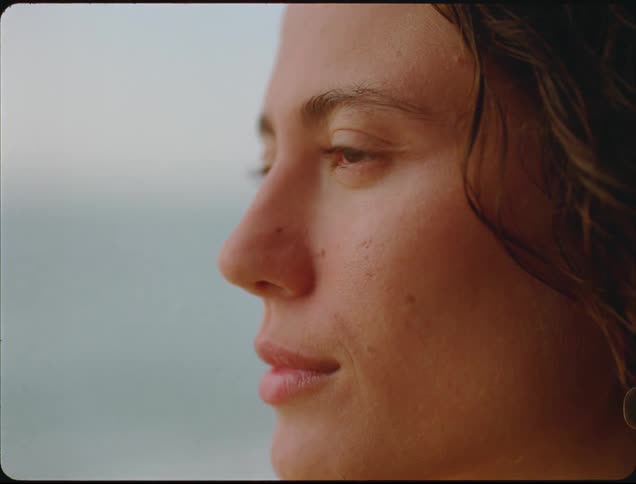 The video is a closeup profile shot of a young womans face with her gaze directed toward the horizon or a distant objecther expression appears thoughtful or contemplative set against a soft blurred background possibly the ocean or sky at dusk
