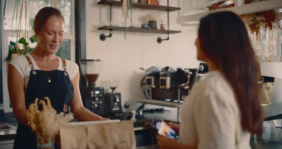 A young woman enters a coffee shop at midday while answering a call sets her phone down and approaches the counter where a sales representative also a young woman steps forward to attend to her needs