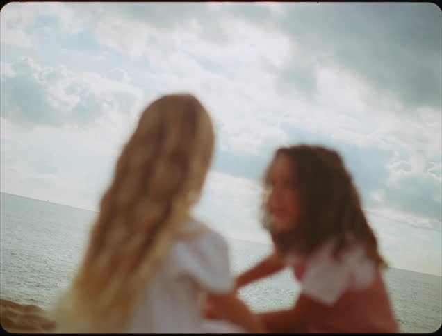 A close view of two young girls with long hair sitting and interacting by the ocean under a cloudy sky captured with a nostalgic soft focus effect