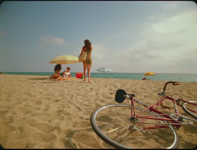 A young lady carrying a handbag walks away from a bicycle lying on the beach sand its back wheel still spinning as she heads toward two people relaxing under an umbrella on the sunny beach