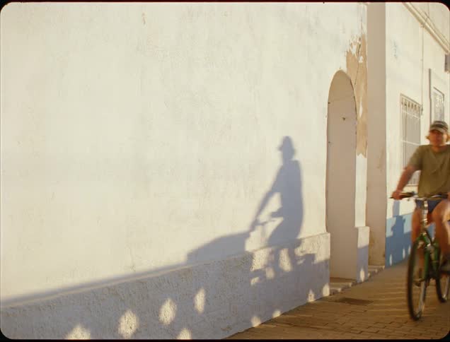 A young man rides his bicycle along the sidewalk on a sunny day wearing a hat