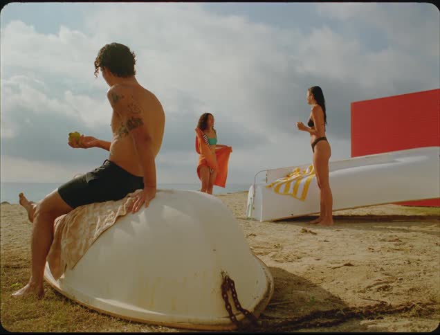 The women are drying off on the beach as a man eats a pear