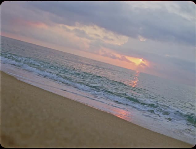 Waves roll into shore and over the beach as the sun shines through clouds on the horizon