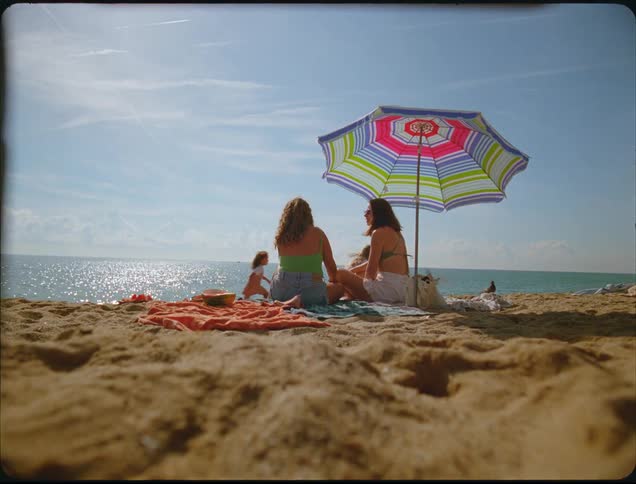 A wide view shows two women relaxing under a colorful umbrella on a sandy beach near the sparkling ocean while young girls run past them