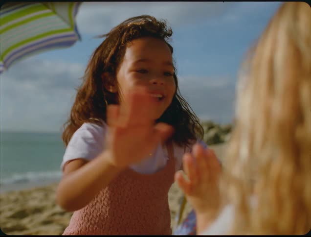 A closeup shot shows a young girl with brown curly hair laughing and happily playing a handclapping game with another person seen only from the back on a sunny beach the scene has a warm vintage film aesthetic with a colorful beach umbrella visibl