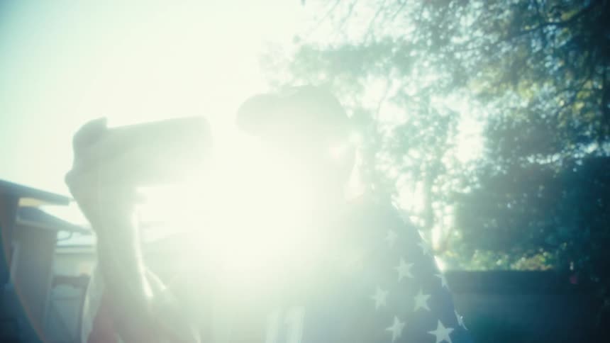 A senior man wearing a baseball cap and a patriotic outfit that mixes a usa flag design with a soccer jersey holds up a smartphone to record or watch something he appears excited and animated cheering enthusiastically outdoors on a very sunny day wit
