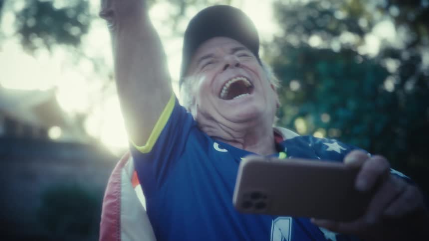 Elderly man wearing a soccer jersey cheering and laughing while looking at a smartphone in his hand outdoors during day time