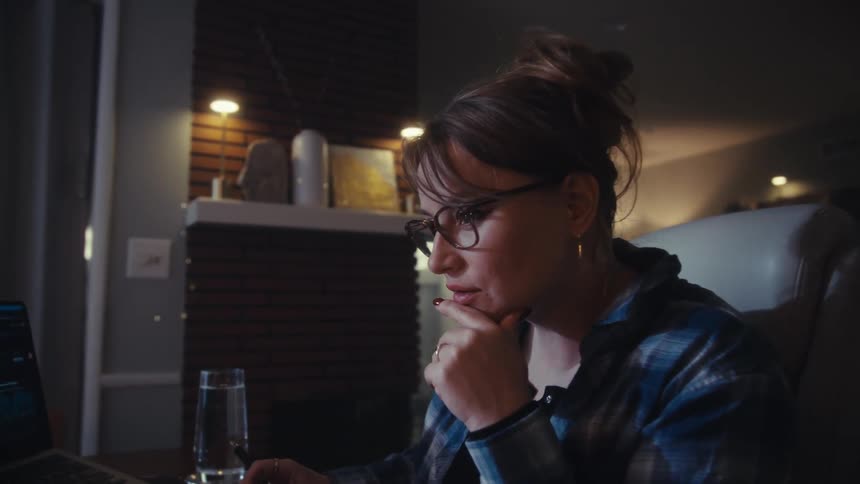 Person with glasses and plaid shirt gesturing in front of laptop in indoor setting with soft lighting