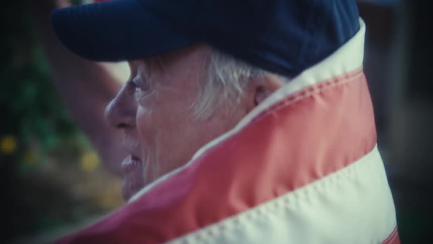 The video shows an older person wearing a baseball cap and draped in an american flag first holding a smartphone and then bringing a whistle or noisemaker to their mouth the scene quickly cuts to a closeup of the person cheering and shouting enthusias