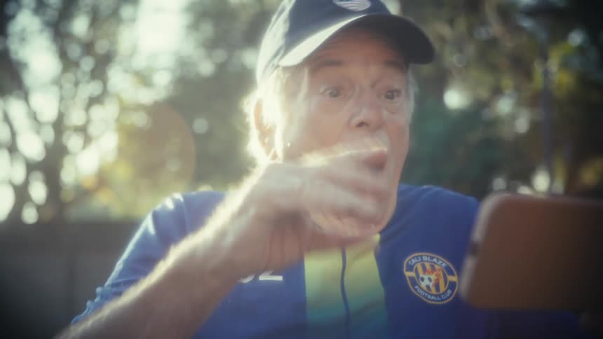An elderly man wearing a cbz jersey and a hat stands in his backyard on a sunny day watching a sports event on his mobile phone and becoming overly excited