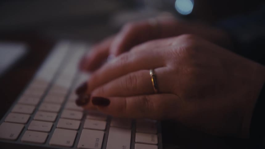 A close view shows a womans hands with rings and painted nails quickly typing on a white keyboard against a dark screen displaying code