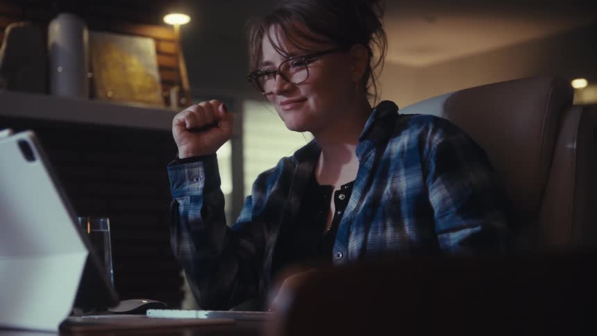 A young woman sits indoors behind a desk at midday wearing glasses watching content on a tablet placed in front of her with a glass of water at her side as she watches in excitement
