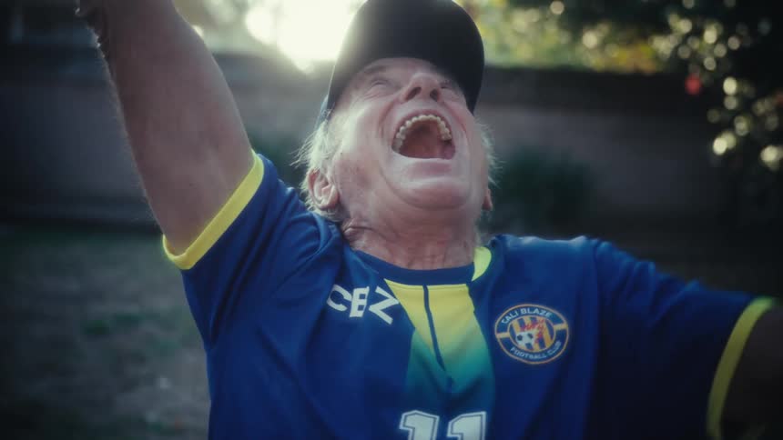 An elderly man wearing a blue and yellow soccer jersey and a dark baseball cap initially holds a smartphone and then raises his arms shouting with an expression of intense joy and excitement while outdoors in a sunny backyard setting