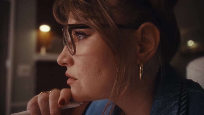 A middleaged woman sits in an office wearing gold earrings glasses and gold rings on her fingers quietly contemplating