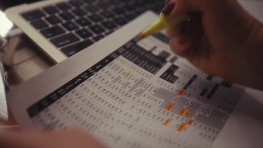 Hand highlighting spreadsheet data with yellow marker next to laptop keyboard