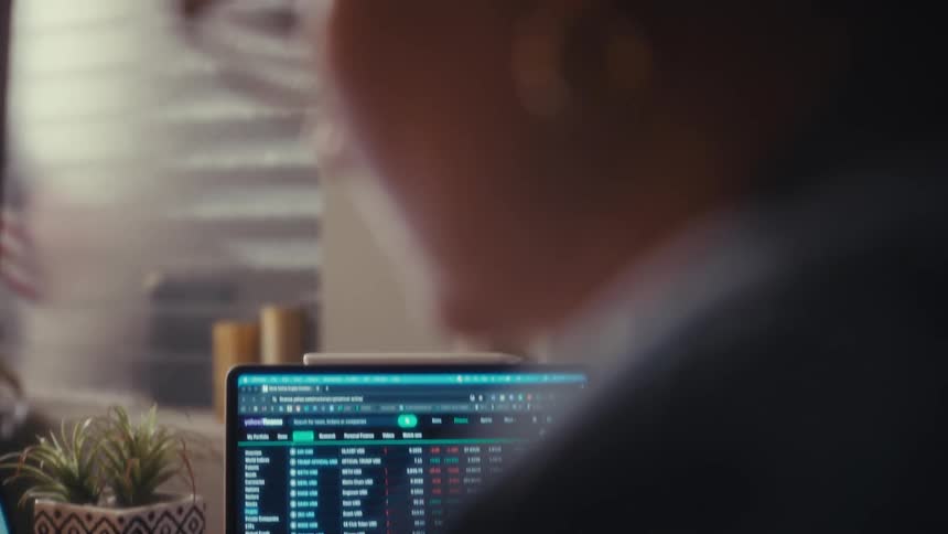 Woman wearing glasses looking at financial information on a laptop and taking down notes on a piece of paper indoors during day time
