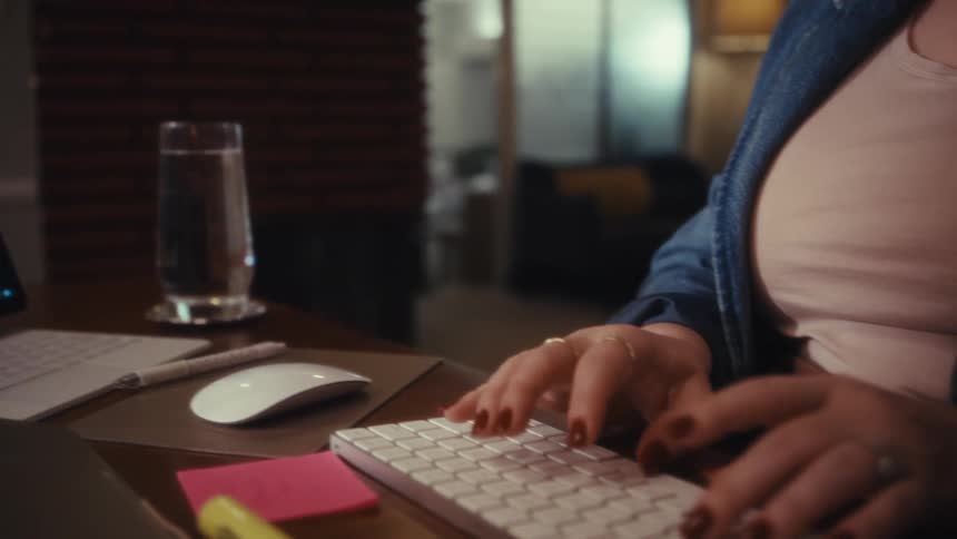 A womans hands with red manicured nails are typing on a white keyboard on a wooden desk next to a glass of water