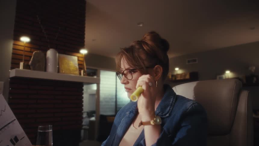 A close view shows a woman wearing glasses and a denim jacket carefully studying a printed chart while working in a dimly lit home office