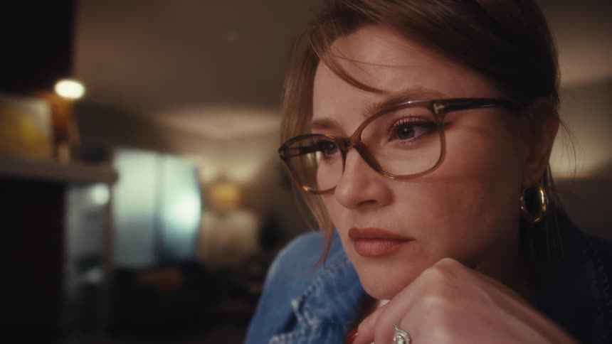 A woman wearing glasses a denim jacket and a watch is shown focusing intently on a screen resting her chin on her hand while holding a pen the camera then shifts focus to a pair of laptops on a desk displaying complex code or financial data in a dimly